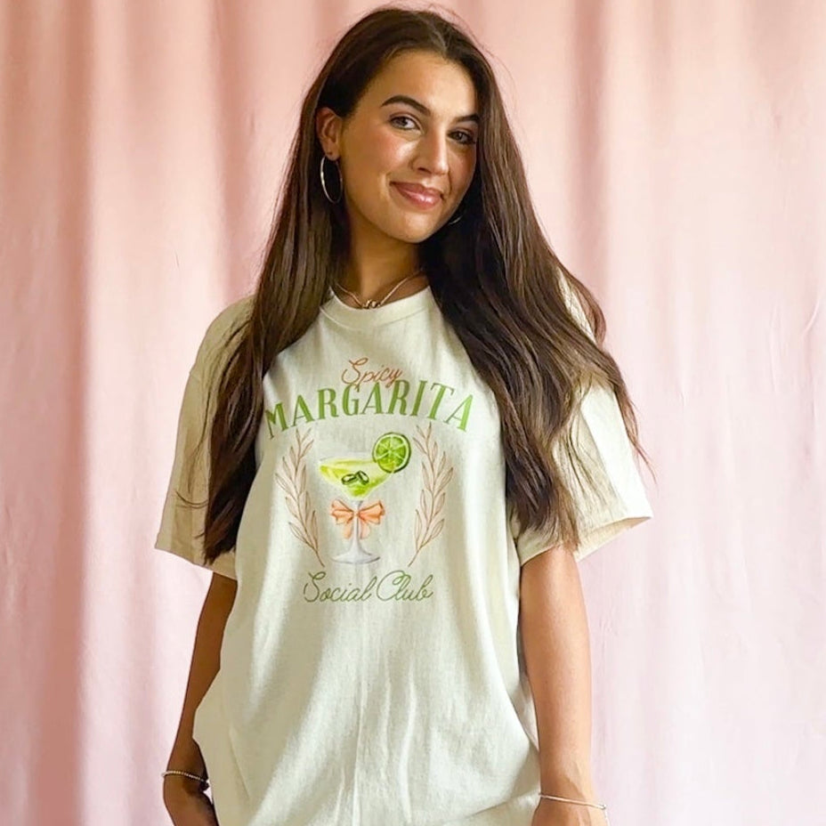 Woman wearing a 'Savvy Margarita Social Club' t-shirt against a pink curtain background