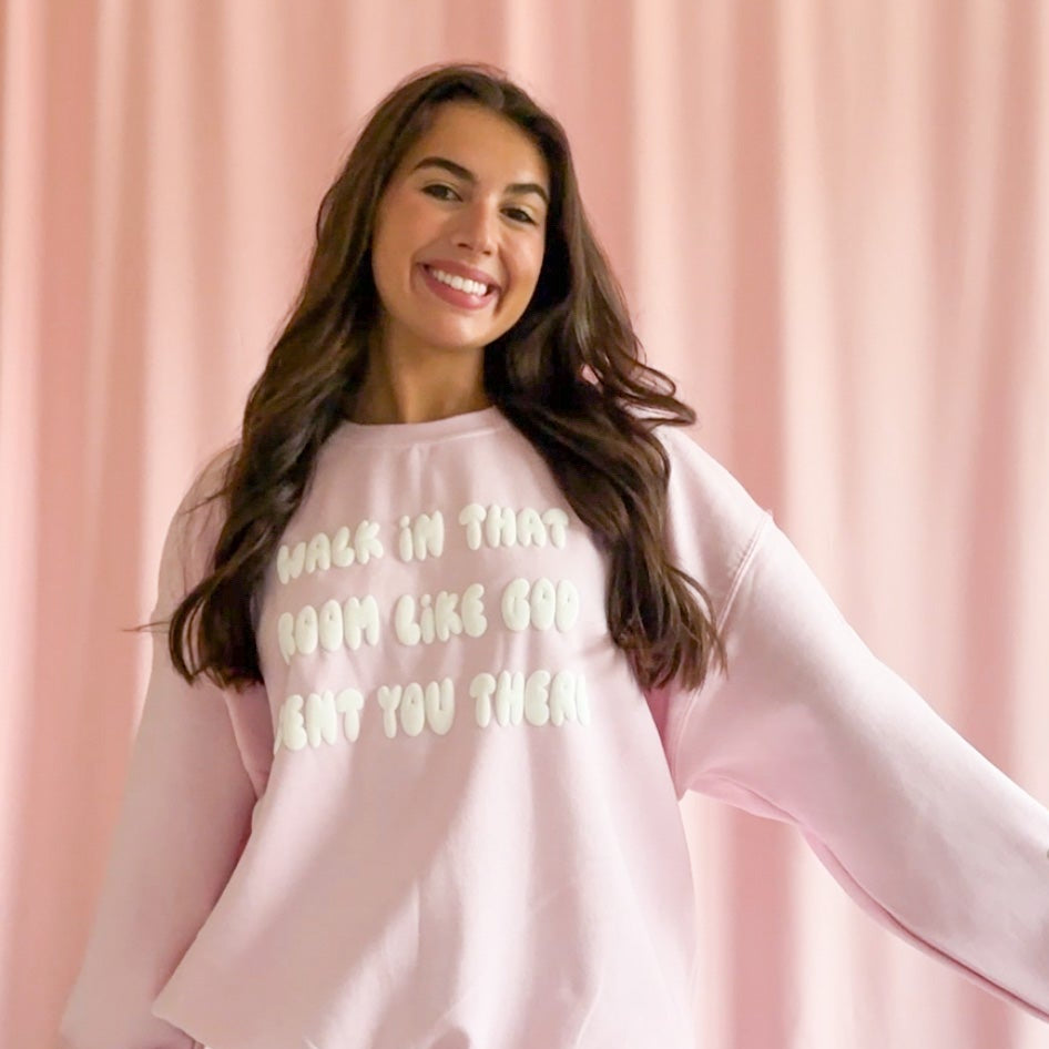 A woman standing in front of a pink backdrop, wearing a pink sweatshirt with the phrase 'Walk In That Room Like God Sent You There' on it, paired with ripped jeans.